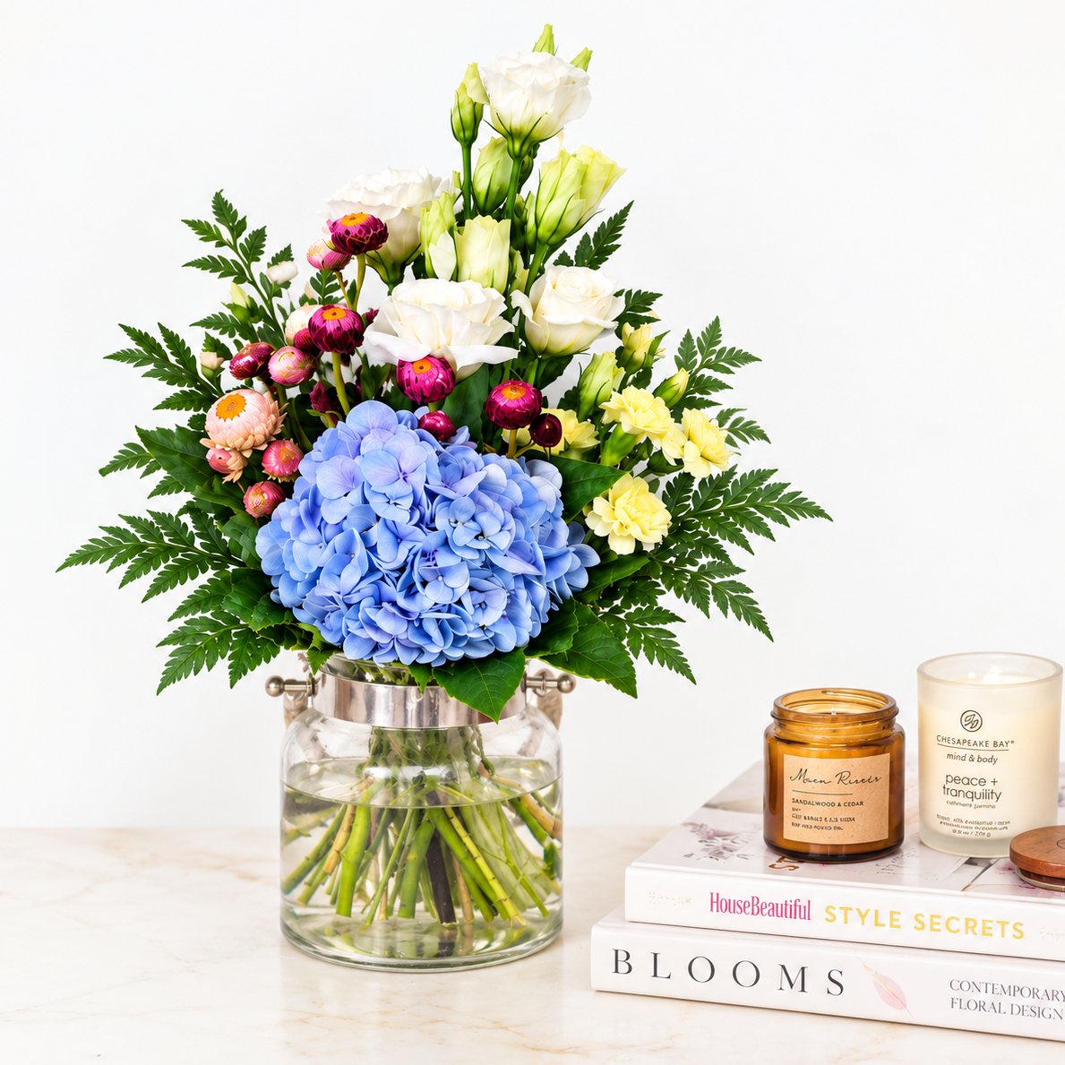 A floral arrangement in a clear glass jar vase with green foliage, blue hydrangea, white Euston, Pink Helichrysum
and yellow Spray Carnation on a marble surface next to two books and two scented candle jars