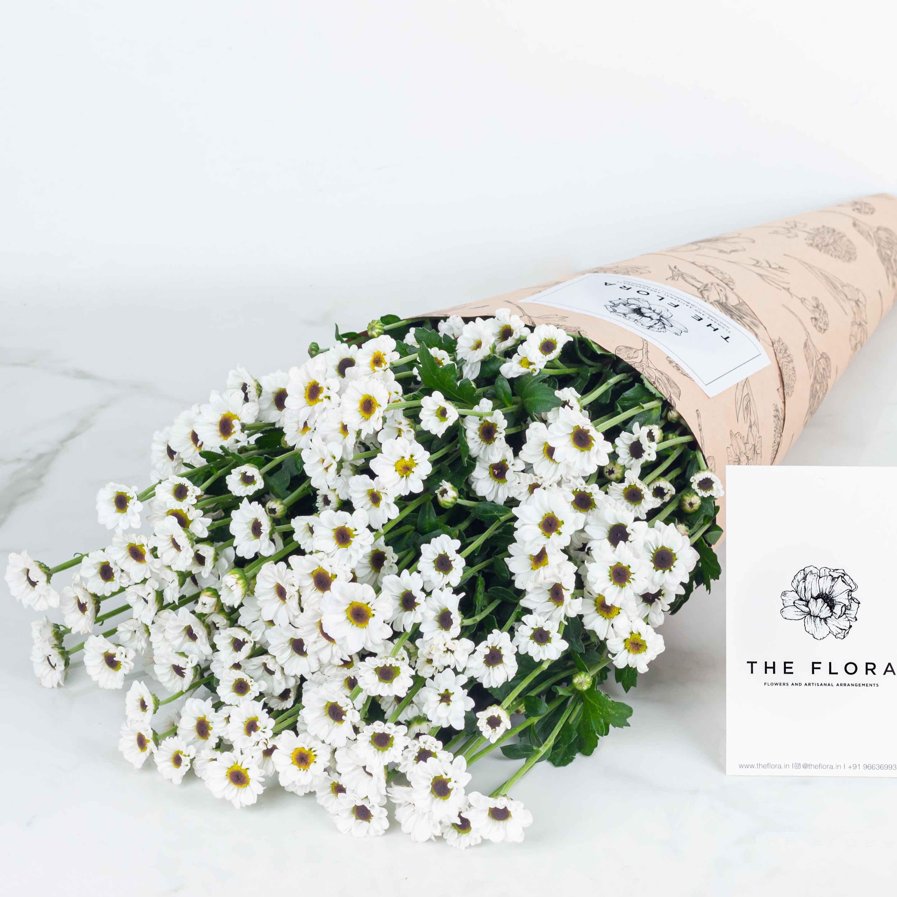 Bouquet of white flowers Santini Chrysanthemum with paper wrap on a light background with 'The Flora' label. Online cut flowers n sustainable packaging from www.theflora.in