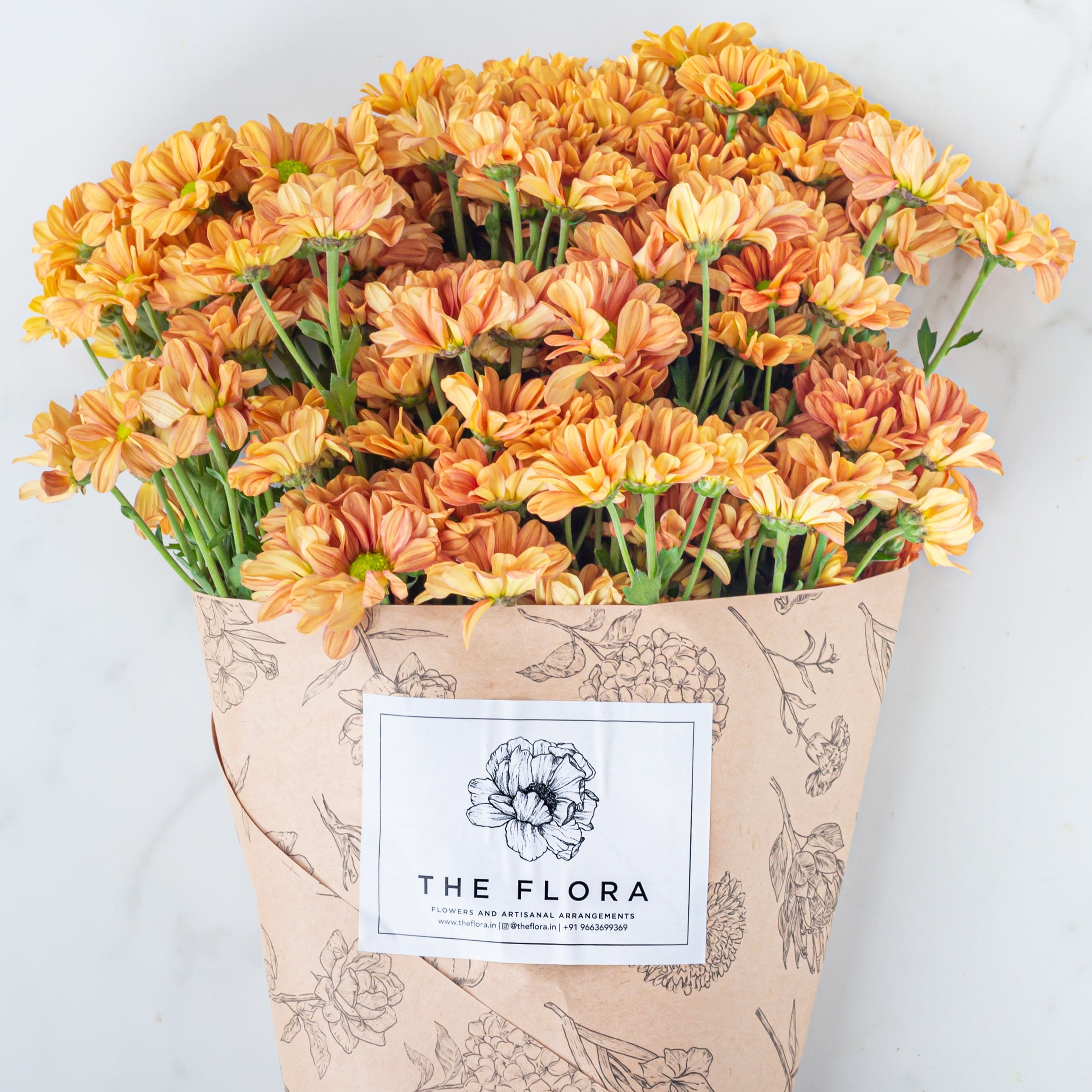 Bouquet of orange flowers Santini Chrysanthemum wrapped in paper with 'The Flora' label . Online cut flowers with same day delivery at www.theflora.in