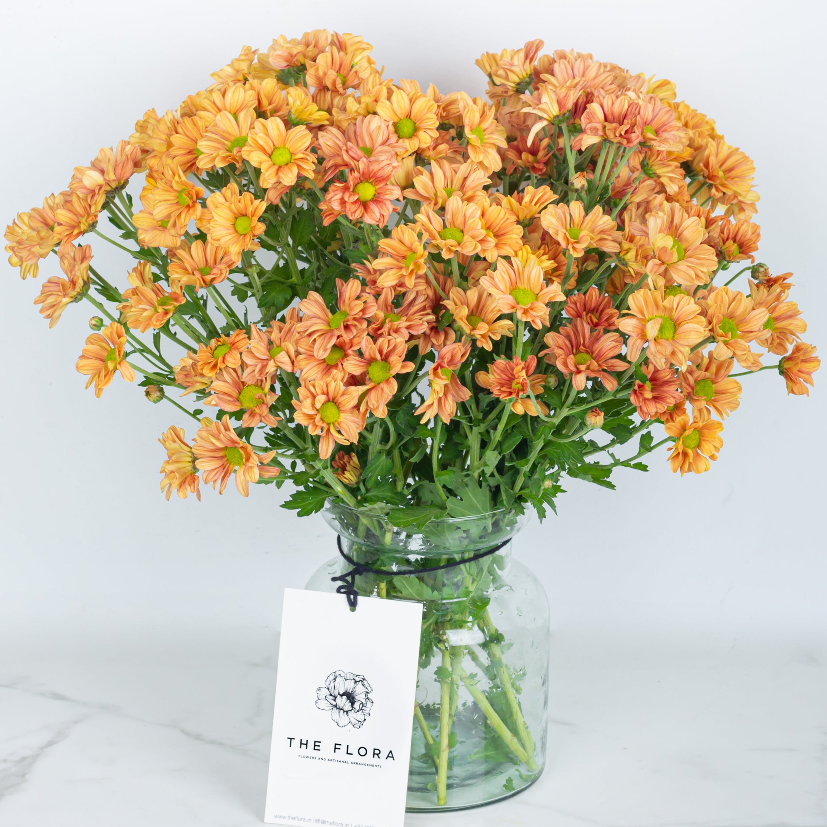 Bouquet of orange Santini Chrysanthemums in a clear vase with 'The Flora' label Online cut flowers with same day delivery at www.theflora.in