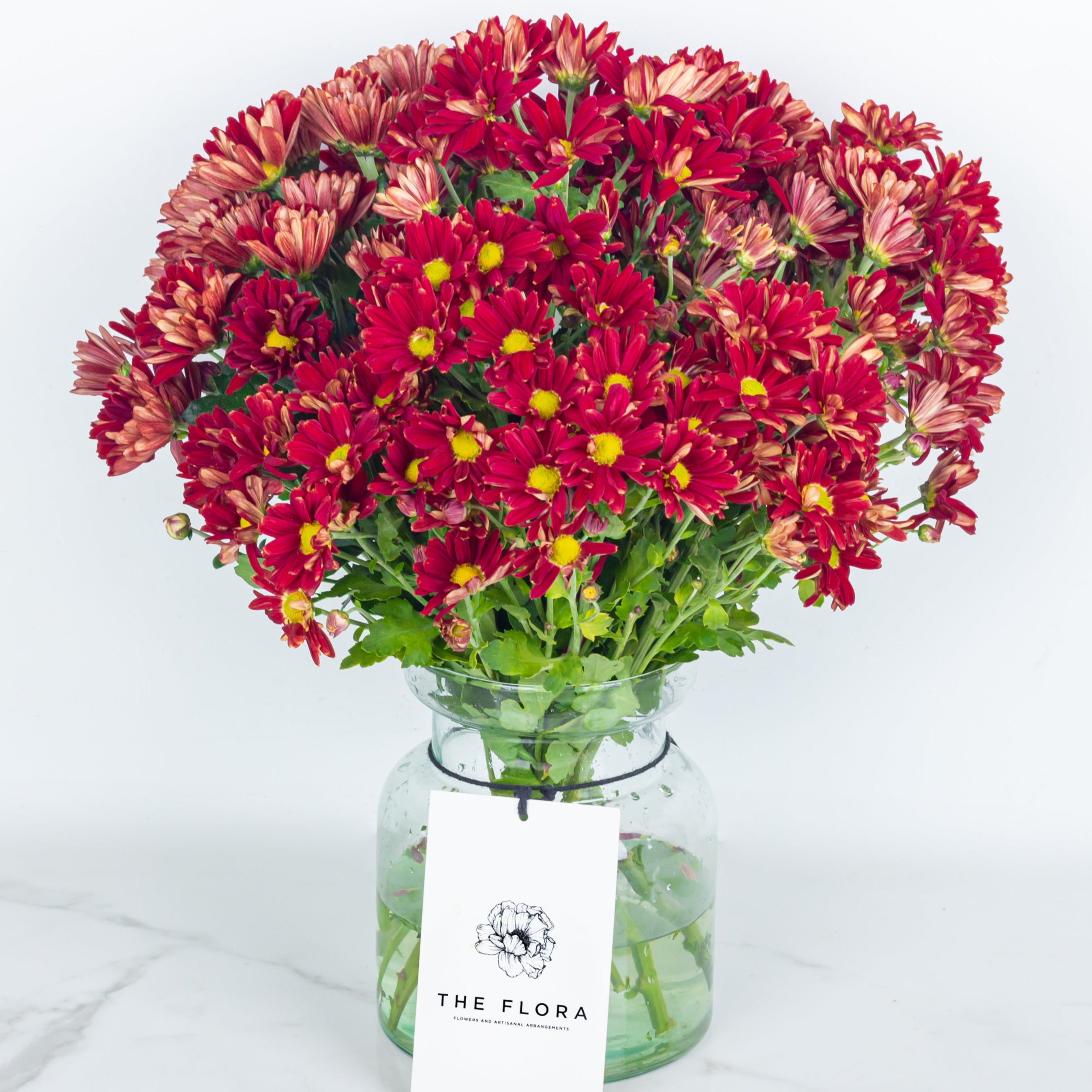 Bouquet of red flowers Santini Chrysanthemums in a glass vase with 'The Flora' tag. Online cut flowers n sustainable packaging from www.theflora.in