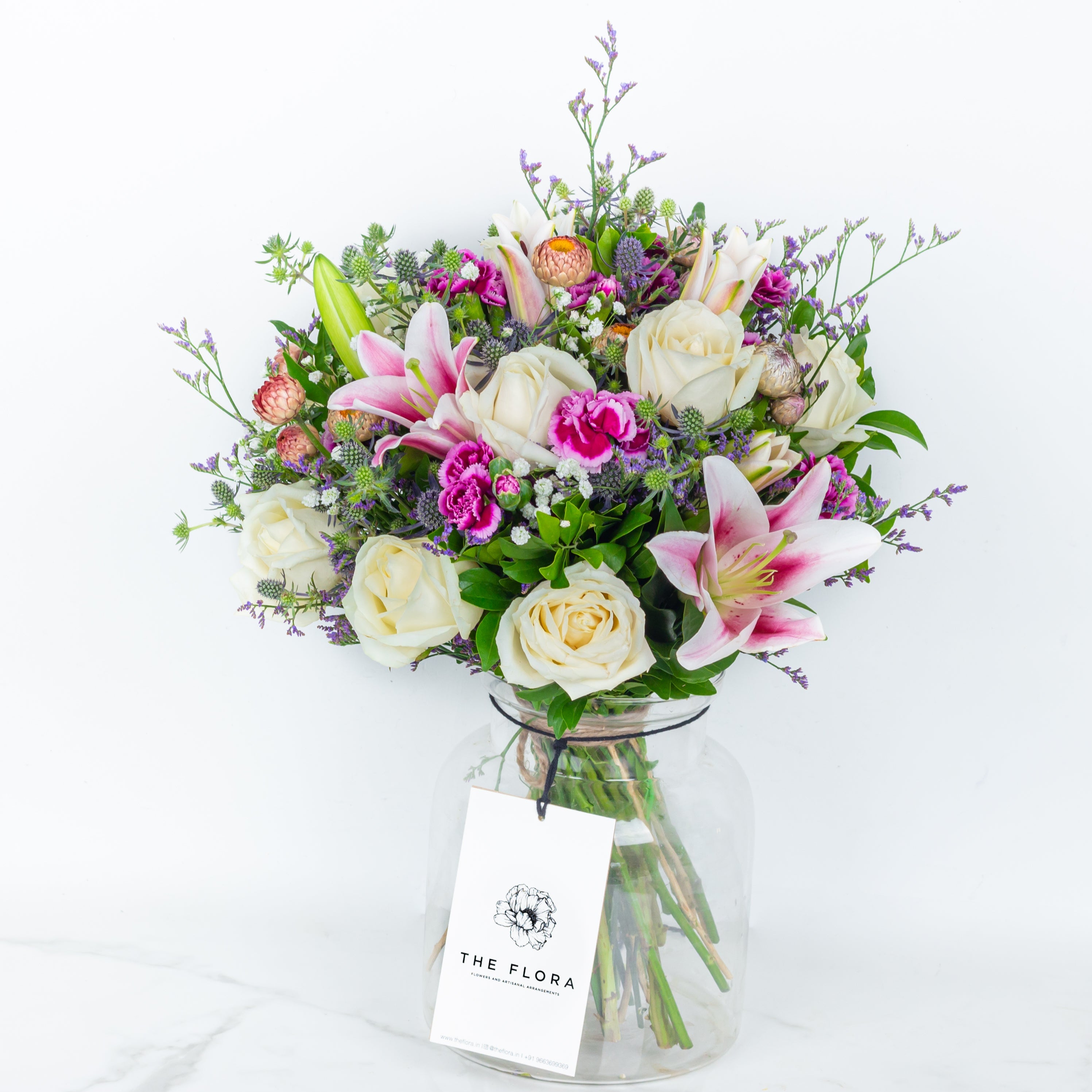 Bouquet of flowers in a clear vase with 'The Flora' label a gift called Soulful Strings . An artisanal bouquet of Eryngium, Roses, Oriental Lilies Helichrysum, Spray Carnations. and Limonium. Online artisanal bouquets with same day delivery at www.theflora.in