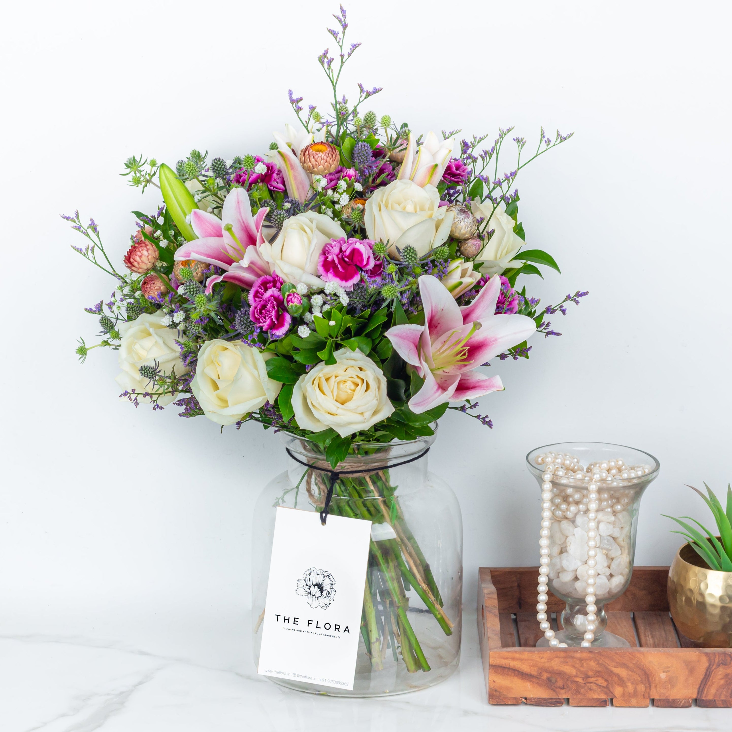 Bouquet of flowers in a clear vase with 'The Flora' label a gift called Soulful Strings . An artisanal bouquet of Eryngium, Roses, Oriental Lilies Helichrysum, Spray Carnations. and Limonium. Online artisanal bouquets with same day delivery at www.theflora.in