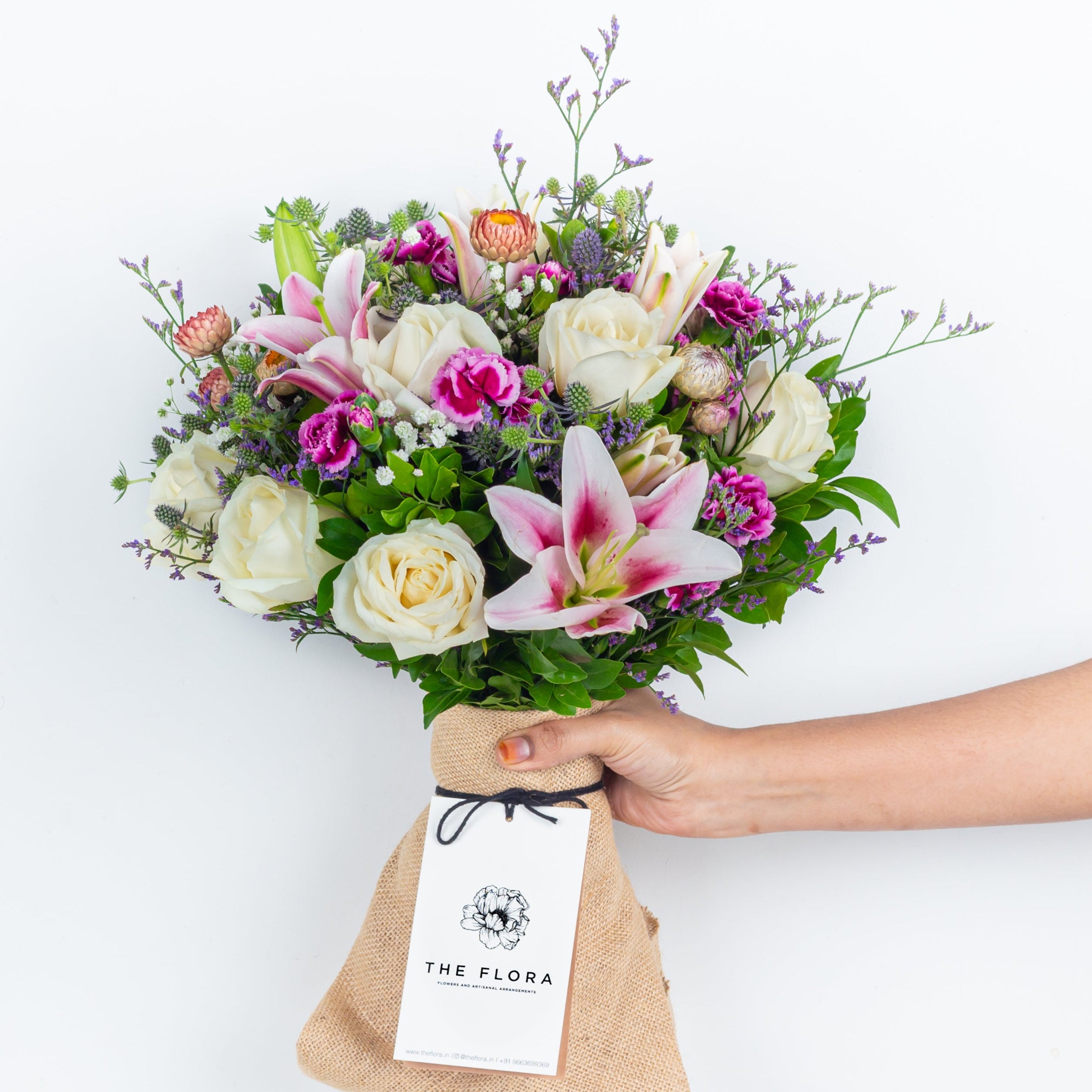 Bouquet of flowers in a clear vase with 'The Flora' label a gift called Soulful Strings . An artisanal bouquet of Eryngium, Roses, Oriental Lilies Helichrysum, Spray Carnations. and Limonium. Online artisanal bouquets with same day delivery at www.theflora.in