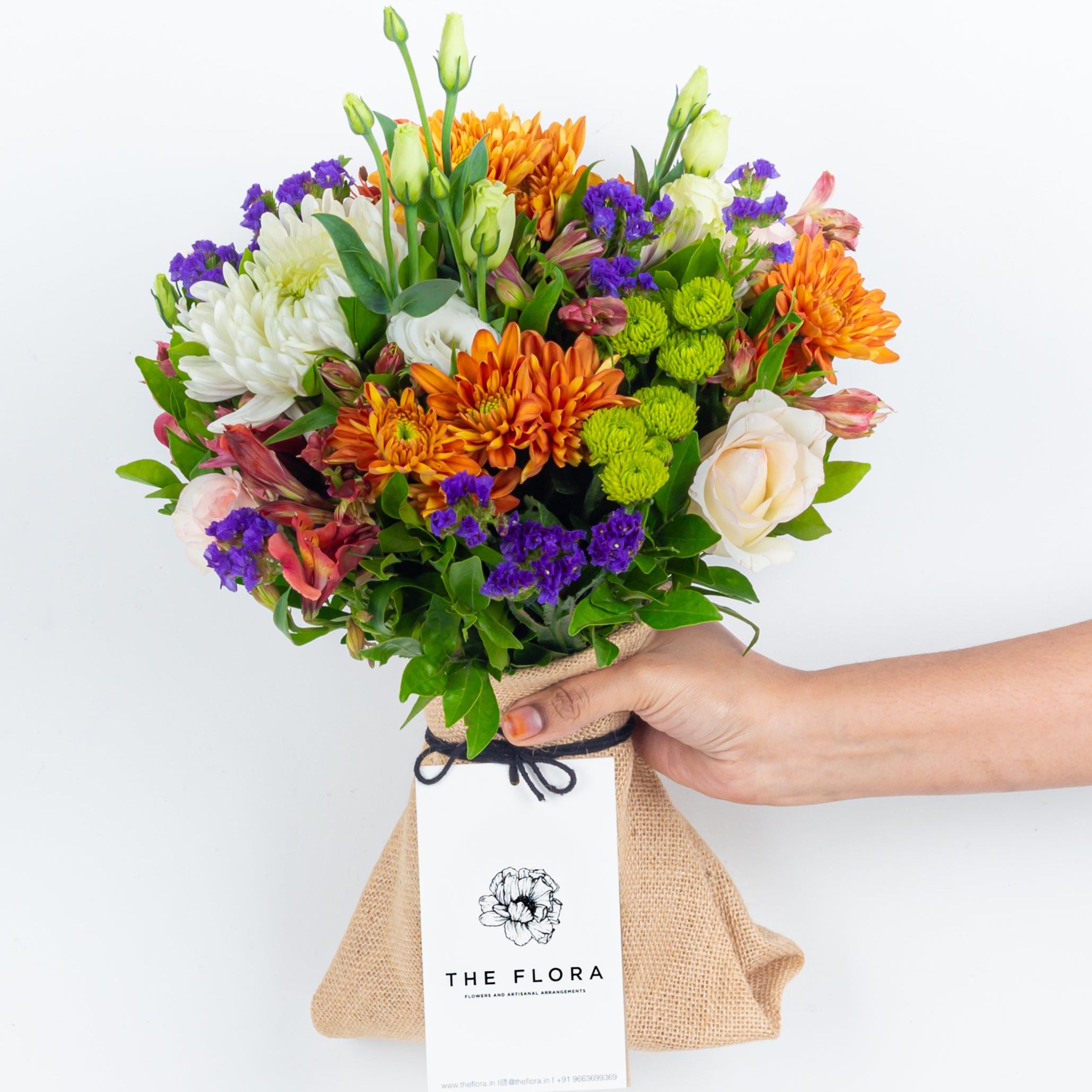 Bouquet of flowers held by a hand with 'The Flora' branding called Guardian Grace with Alstroemeria, Lisianthus , Disbud Chrysanthemums, Blue Statice and Roses. Online artisanal bouquets with same day delivery in sustainable packaging at www.theflora.in