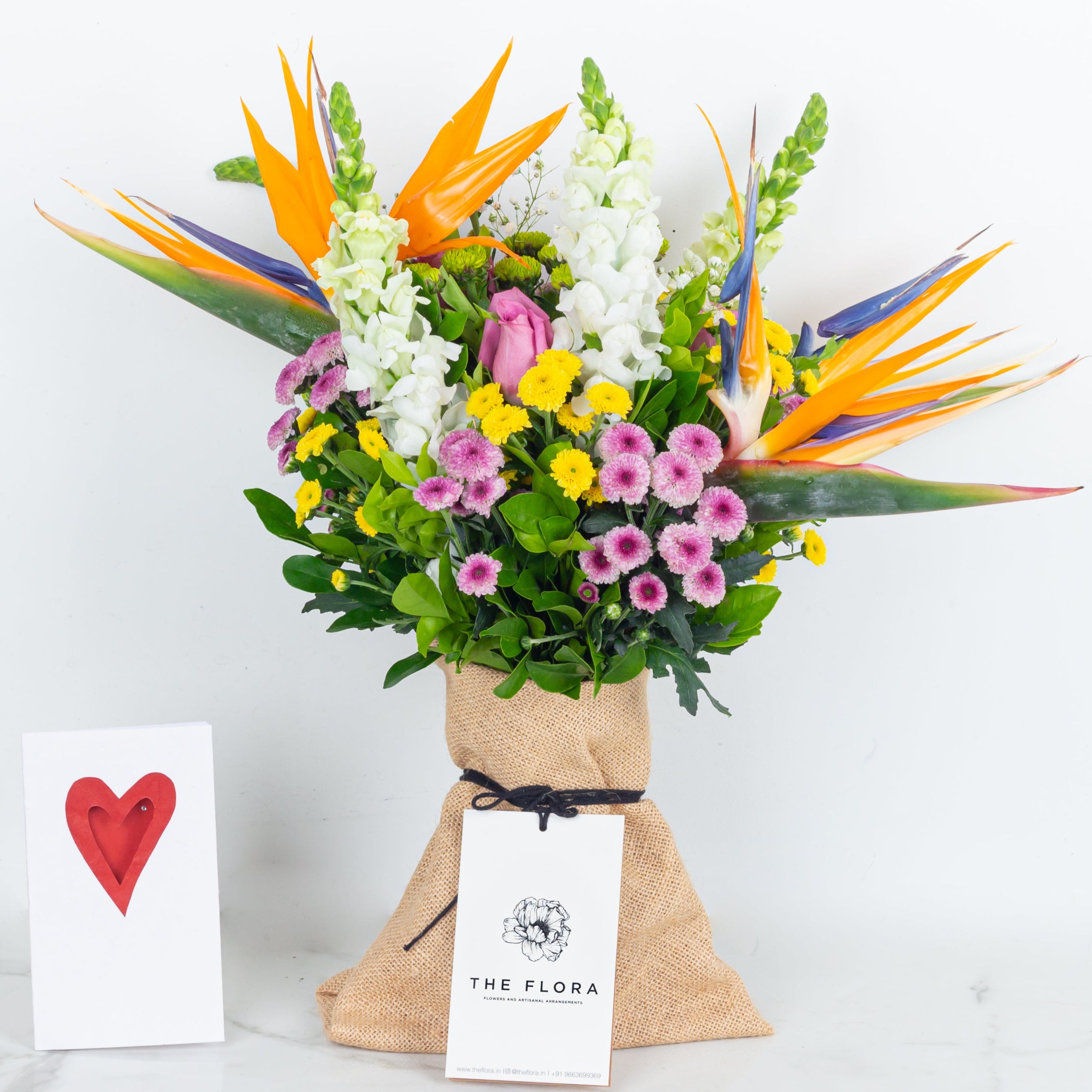 Colorful bouquet of flowers with a 'The Flora' tag called With All My Heart and a card. An artisanal bouquet curated with Birds of Paradise, Button Chrysanthemums and roses. Online artisanal bouquets with same day delivery in sustainable packaging at www.theflora.in