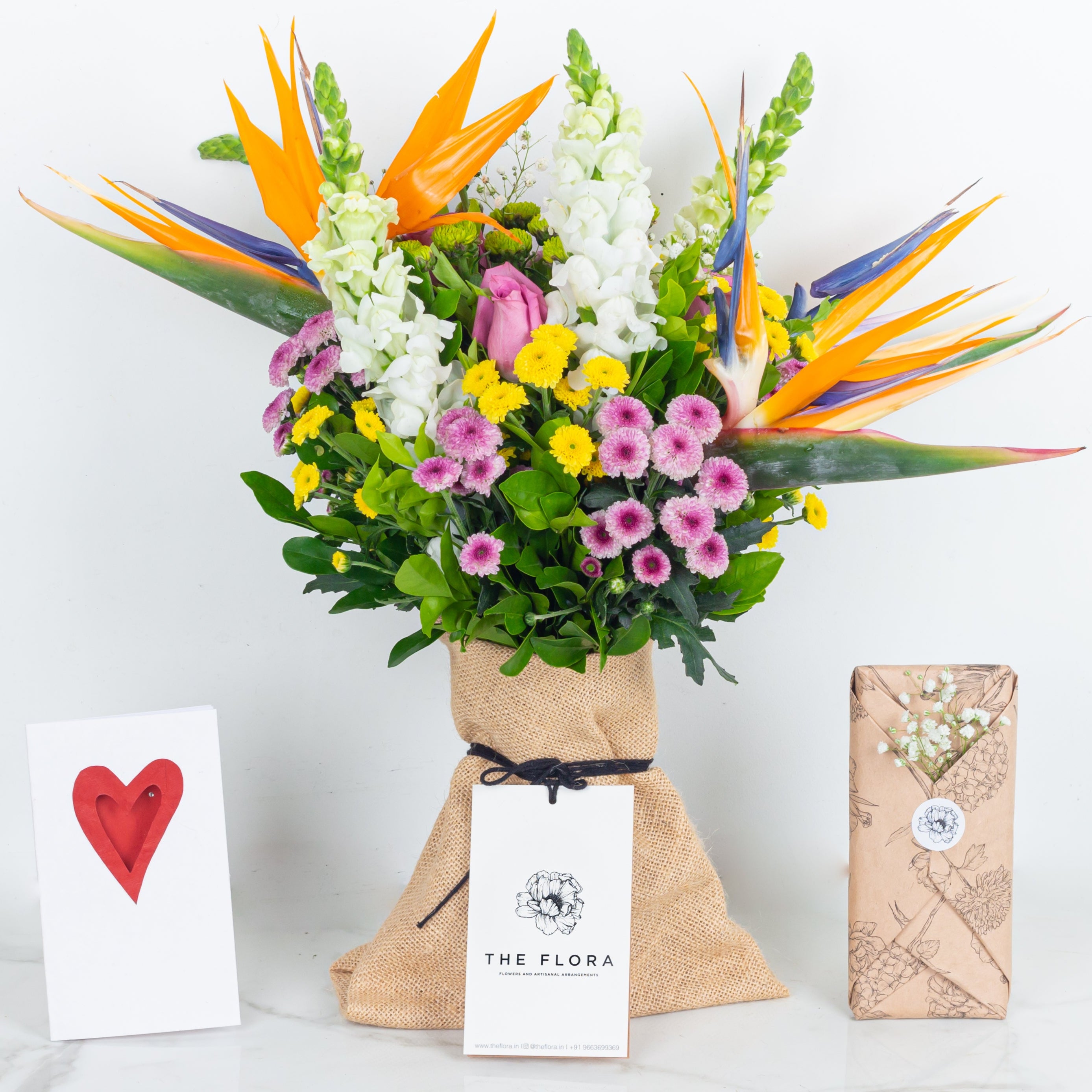 Colorful bouquet of flowers with a 'The Flora' tag called With All My Heart with a card and Chocolate in floral wrapping. An artisanal bouquet curated with Birds of Paradise, Button Chrysanthemums and roses. Online artisanal bouquets with same day delivery in sustainable packaging at www.theflora.in