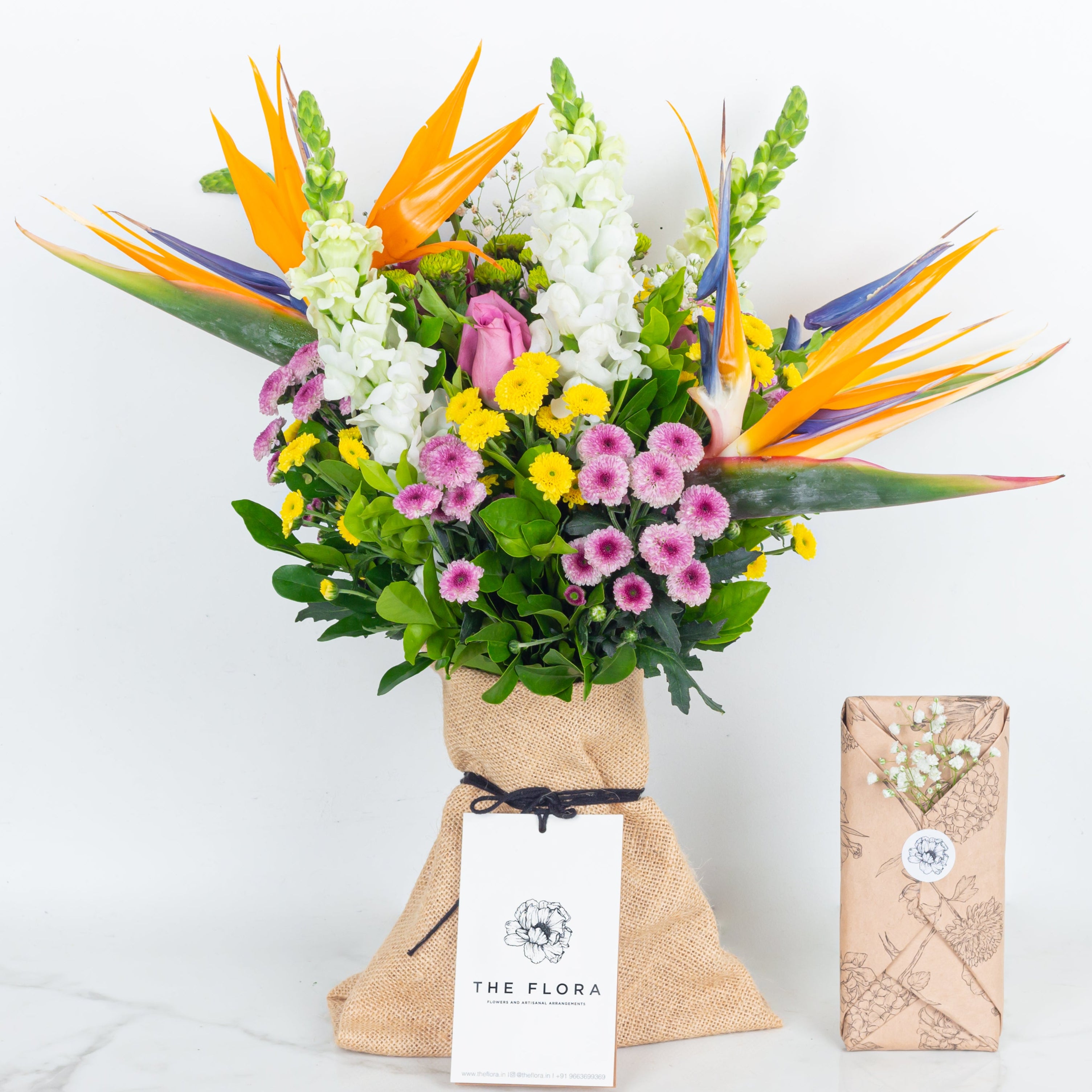 Colorful bouquet of flowers with a 'The Flora' tag called With All My Heart and a Chocolate in floral wrapping. An artisanal bouquet curated with Birds of Paradise, Button Chrysanthemums and roses. Online artisanal bouquets with same day delivery in sustainable packaging at www.theflora.in