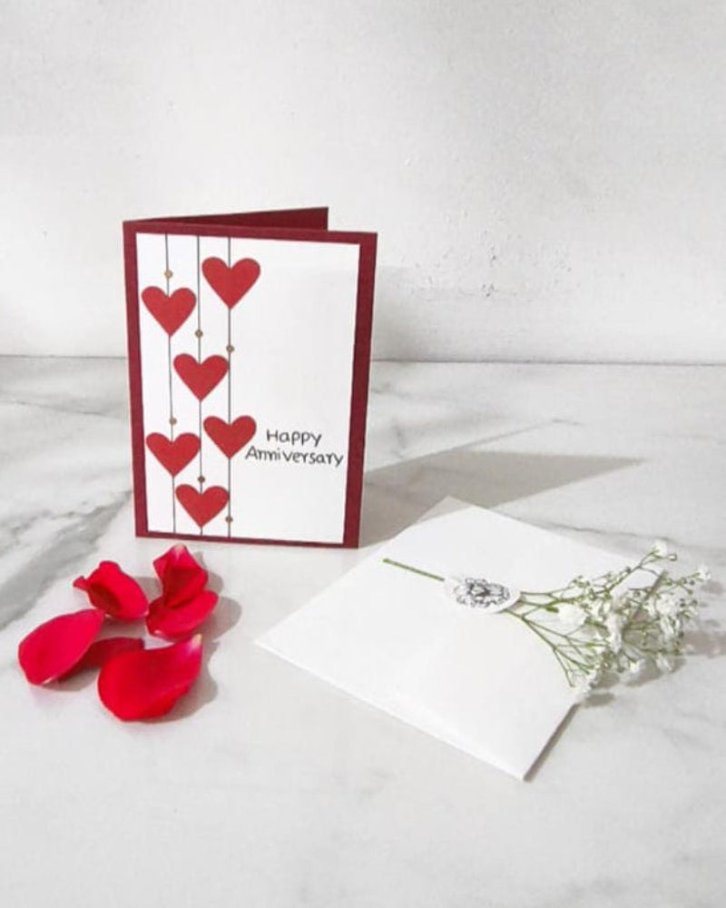 A Happy Anniversary Card with heart designs and red rose petals in a white envelope with a Gypso