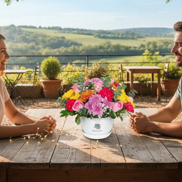 Rainbow Rhapsody basket roses carnations chrysanthemums Bangalore delivery on a table  in a scenic spot with a couple sitting beside it