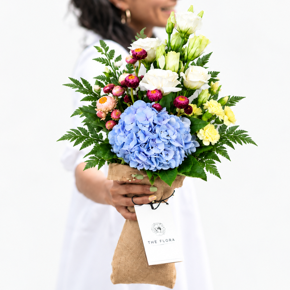 A smiling woman in a white shirt holds a vibrant flower bouquet featuring light blue hydrangeas, white Euston, yellow carnations, and green foliage. The bouquet is wrapped in a brown jute  sleeve with a white card that reads 'THE FLORA'.