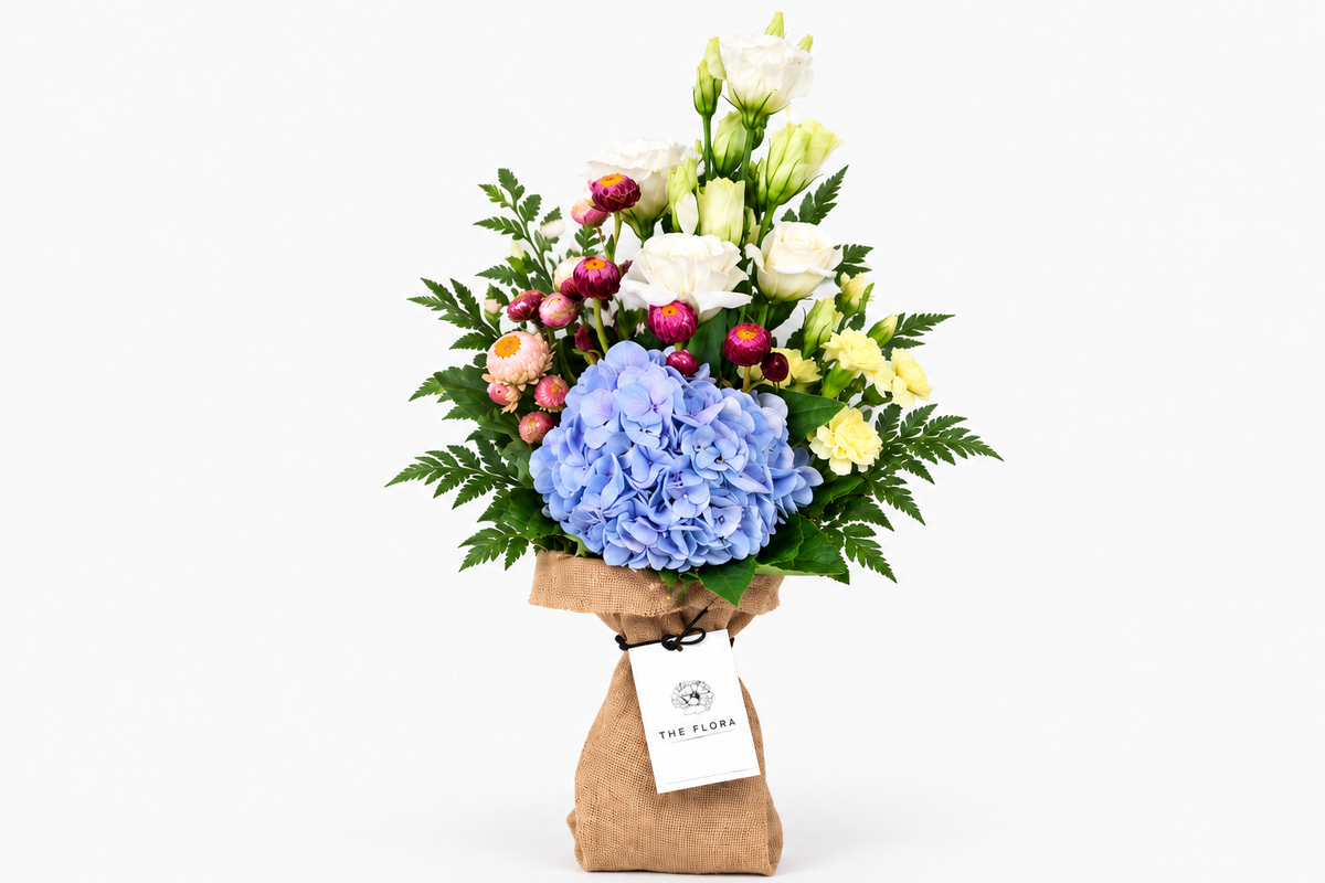 A floral bouquet wrapped in brown jute with a white tag reading 'THE FLORA', featuring a large blue hydrangea, white Eustoma, yellow  Spray Carnation, pink Helicrysum Brown Leather Leaves, and green foliage of Dracaena leaves on a light gray background.




