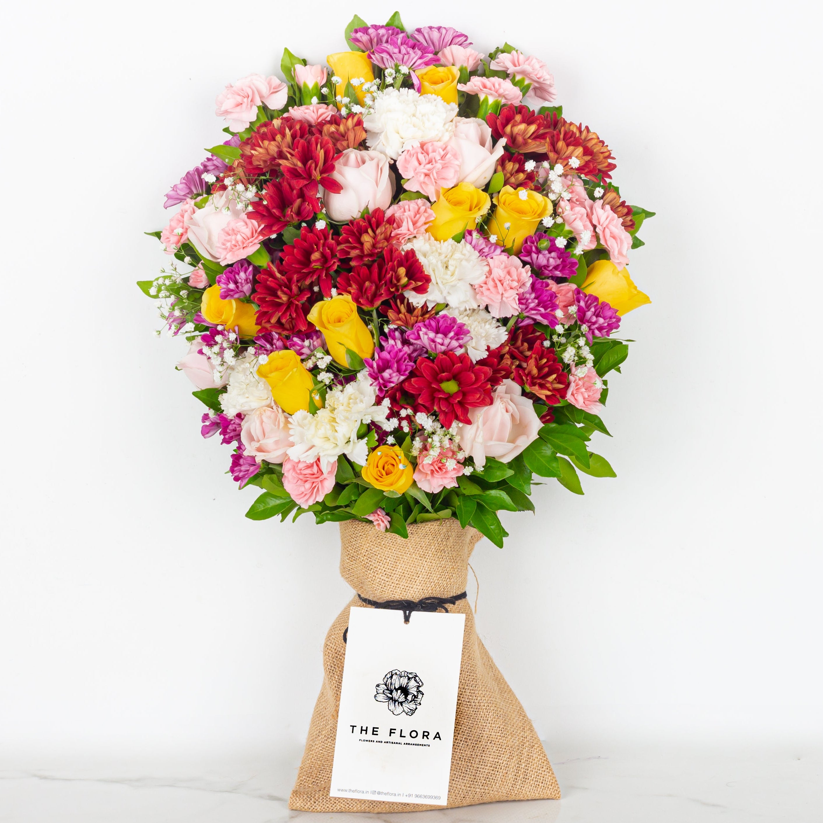 Colorful bouquet of flowers wrapped in brown paper with a white tag on a white background, featuring the brand 'The Flora'.
