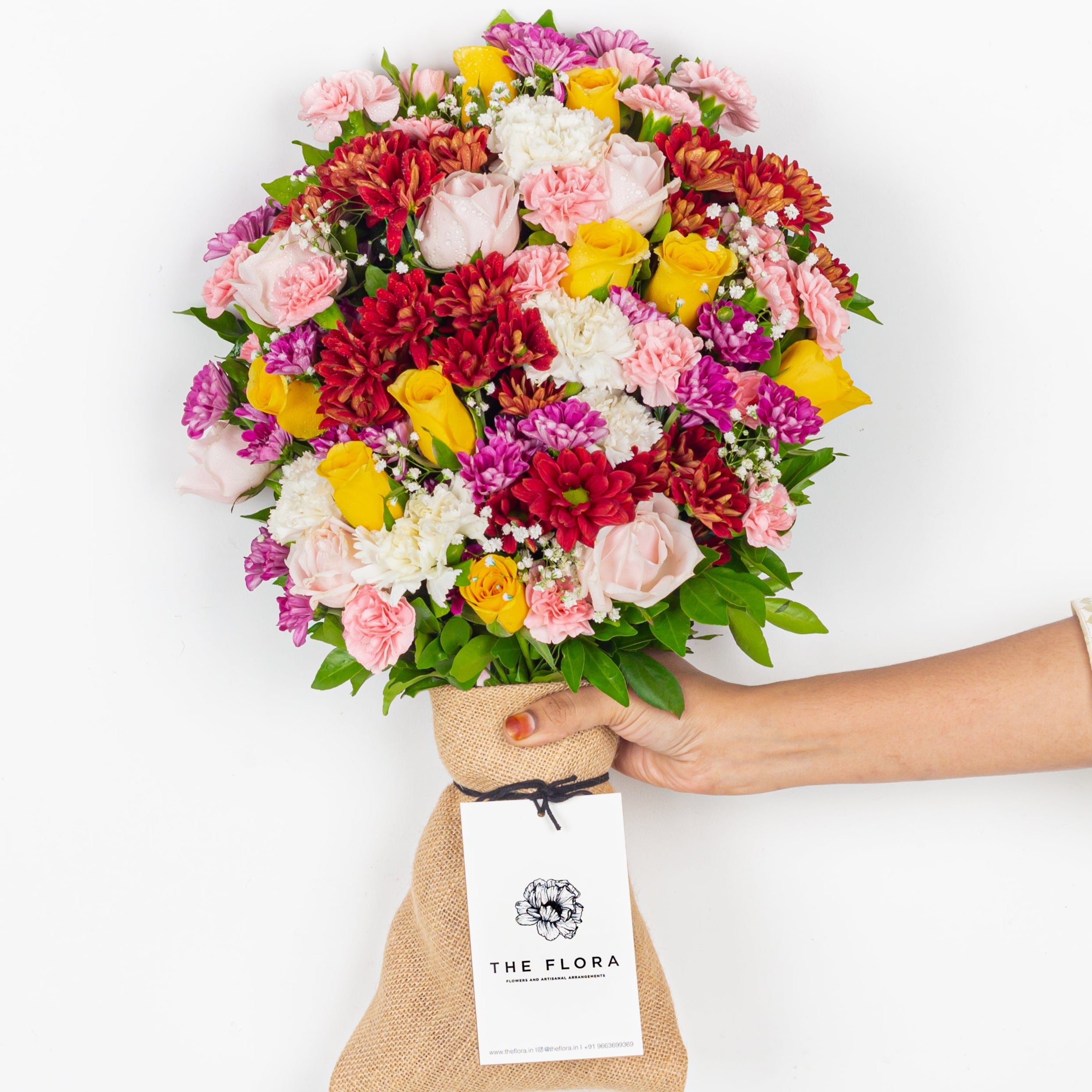 Bouquet of colorful flowers held by a hand with 'The Flora' tag on a white background