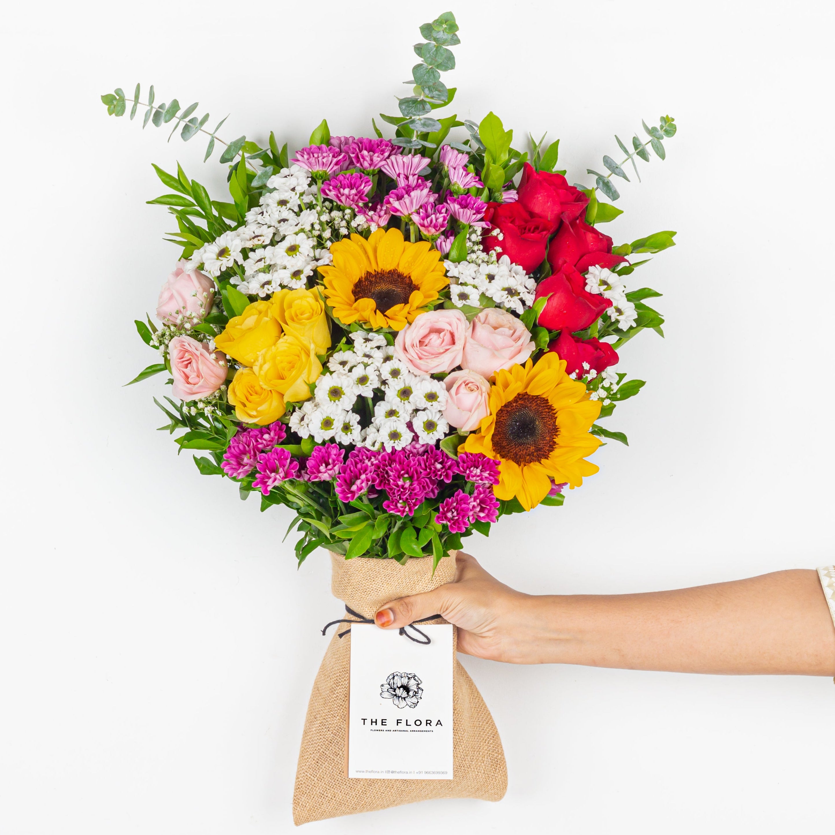 Bouquet of colorful flowers held by a person with 'The Flora' label on a white background