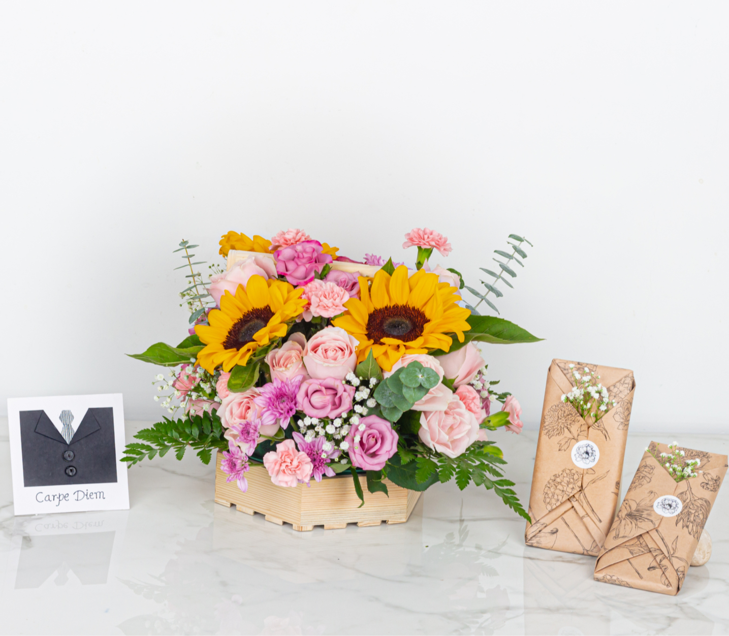 Bouquet of flowers with sunflowers and pink roses, accompanied by two wrapped gifts on a white surface.