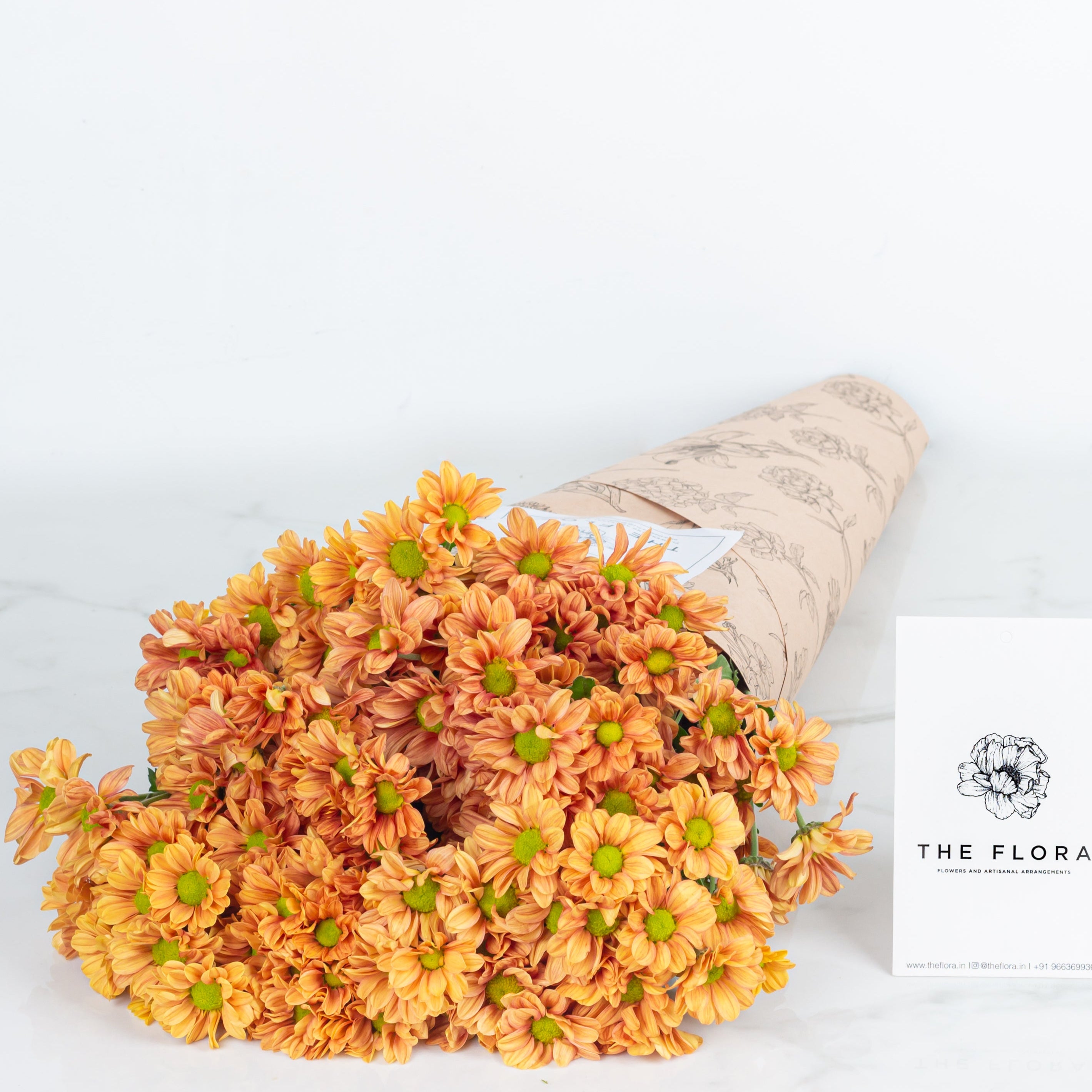 Bouquet of orange flowers Santini Chrysanthemum wrapped in brown paper with 'The Flora' logo. Online cut flowers with same day delivery at www.theflora.in