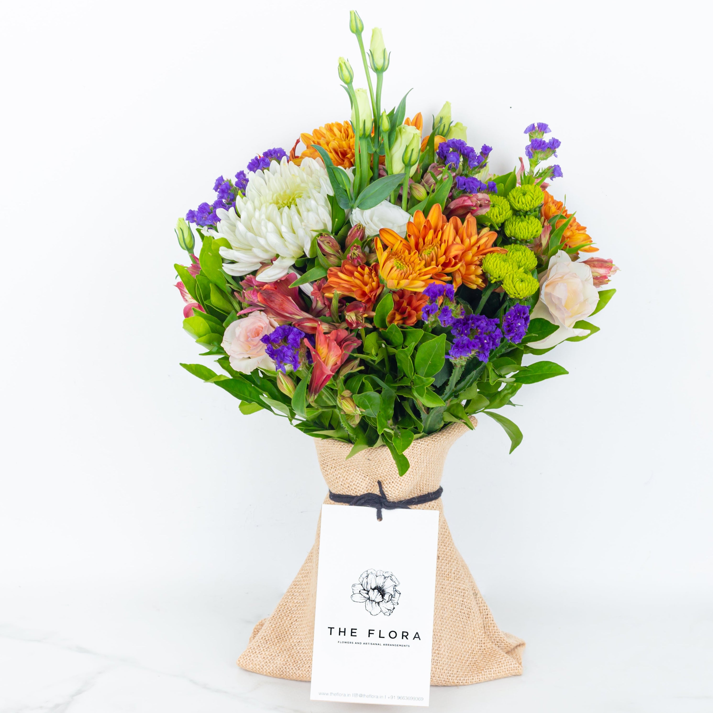 Colorful bouquet of flowers wrapped in paper with 'The Flora' label called Guardian Grace with Alstroemeria, Lisianthus , Disbud Chrysanthemums, Blue Statice and Roses. Online artisanal bouquets with same day delivery in sustainable packaging at www.theflora.in