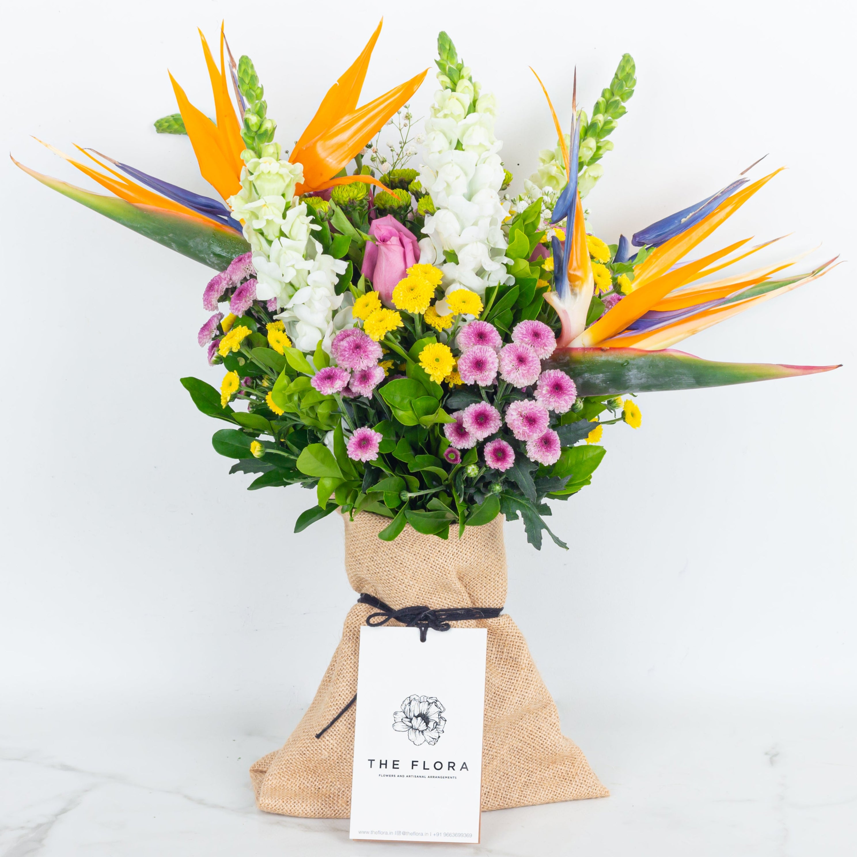 Colorful bouquet of flowers with a 'The Flora' tag called  With All My Heart and a Chocolate in floral wrapping. An artisanal bouquet curated with Birds of Paradise, Button Chrysanthemums and roses. Online artisanal bouquets with same day delivery in sustainable packaging at www.theflora.in