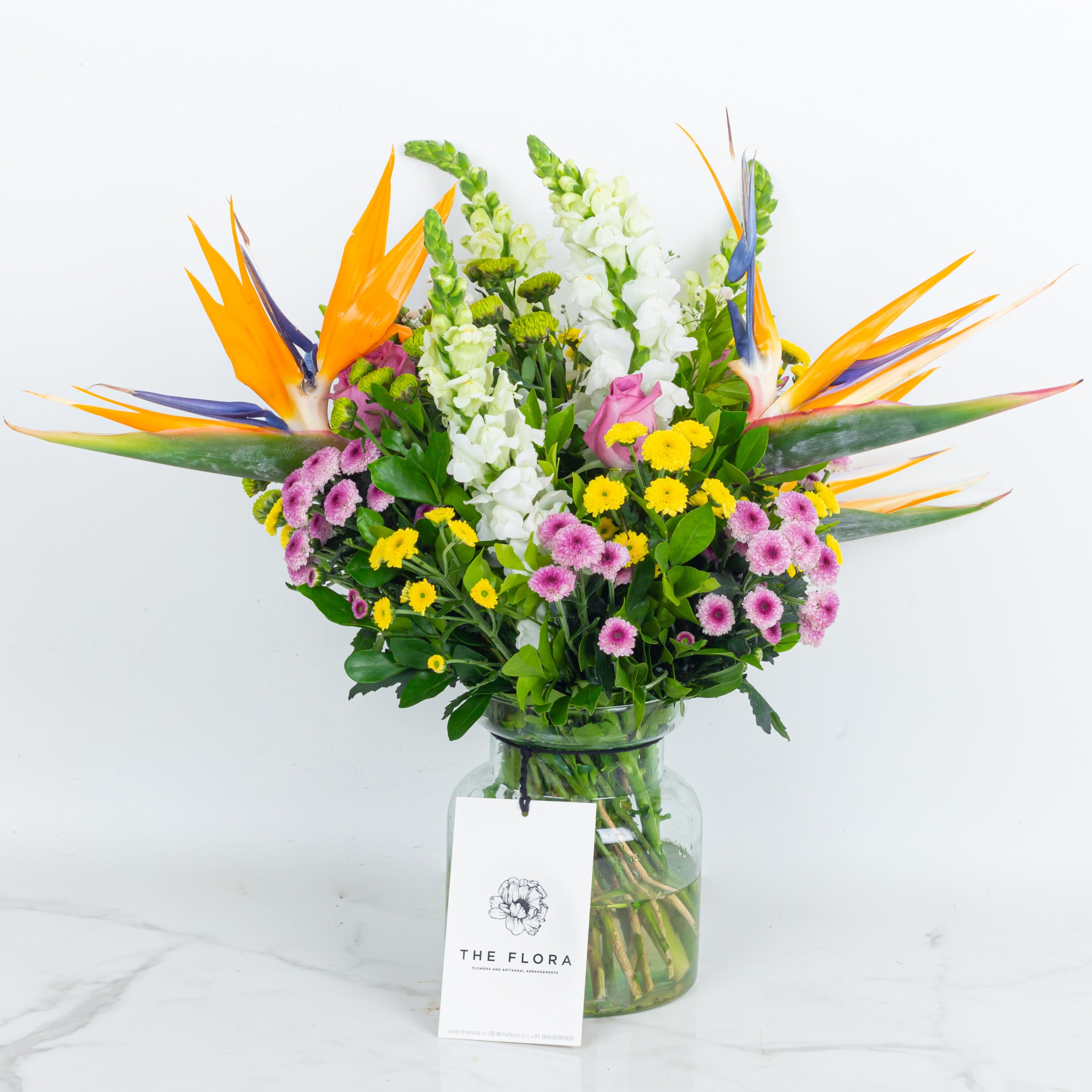 Colorful bouquet of flowers in a glass vase with 'The Flora' label on a white  called with all my heart curated with Birds of Paradise, Button Chrysanthemums and roses. Online artisanal bouquets with same day delivery in sustainable packaging at www.theflora.in
