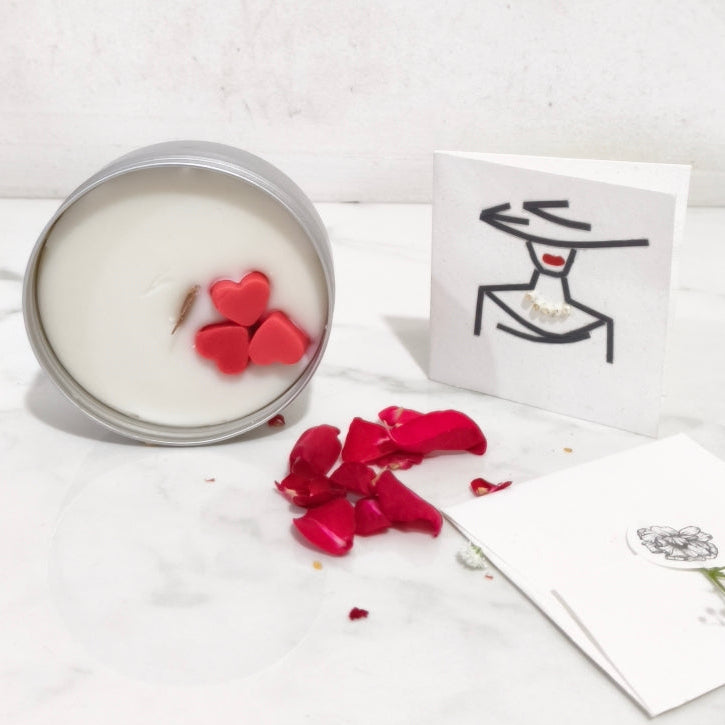 Candle with red heart-shaped wick, red petals, and a card on a white surface