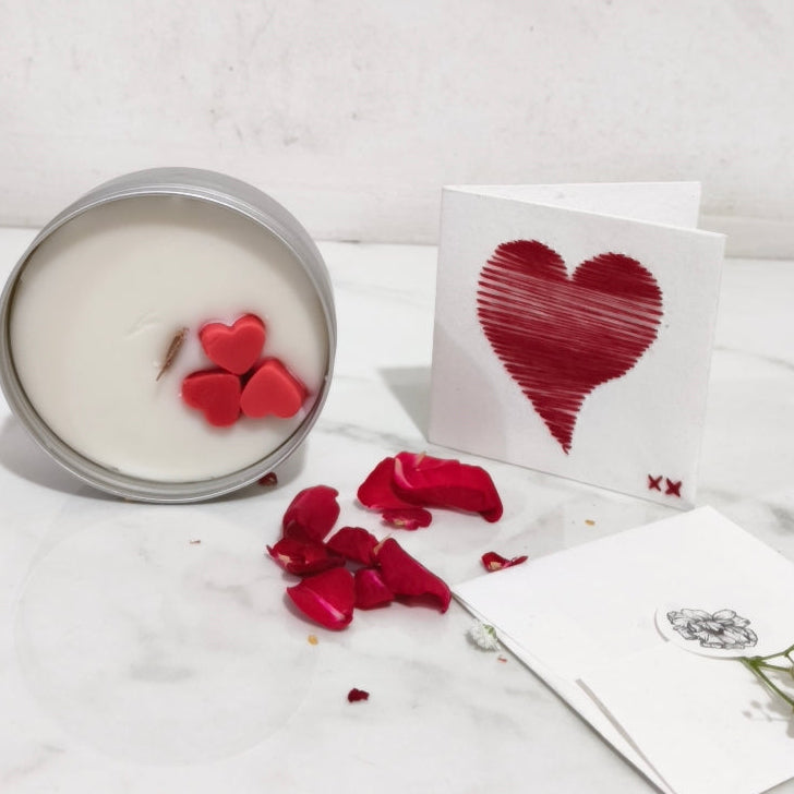 Candle with red heart design, rose petals, and a card on a white surface